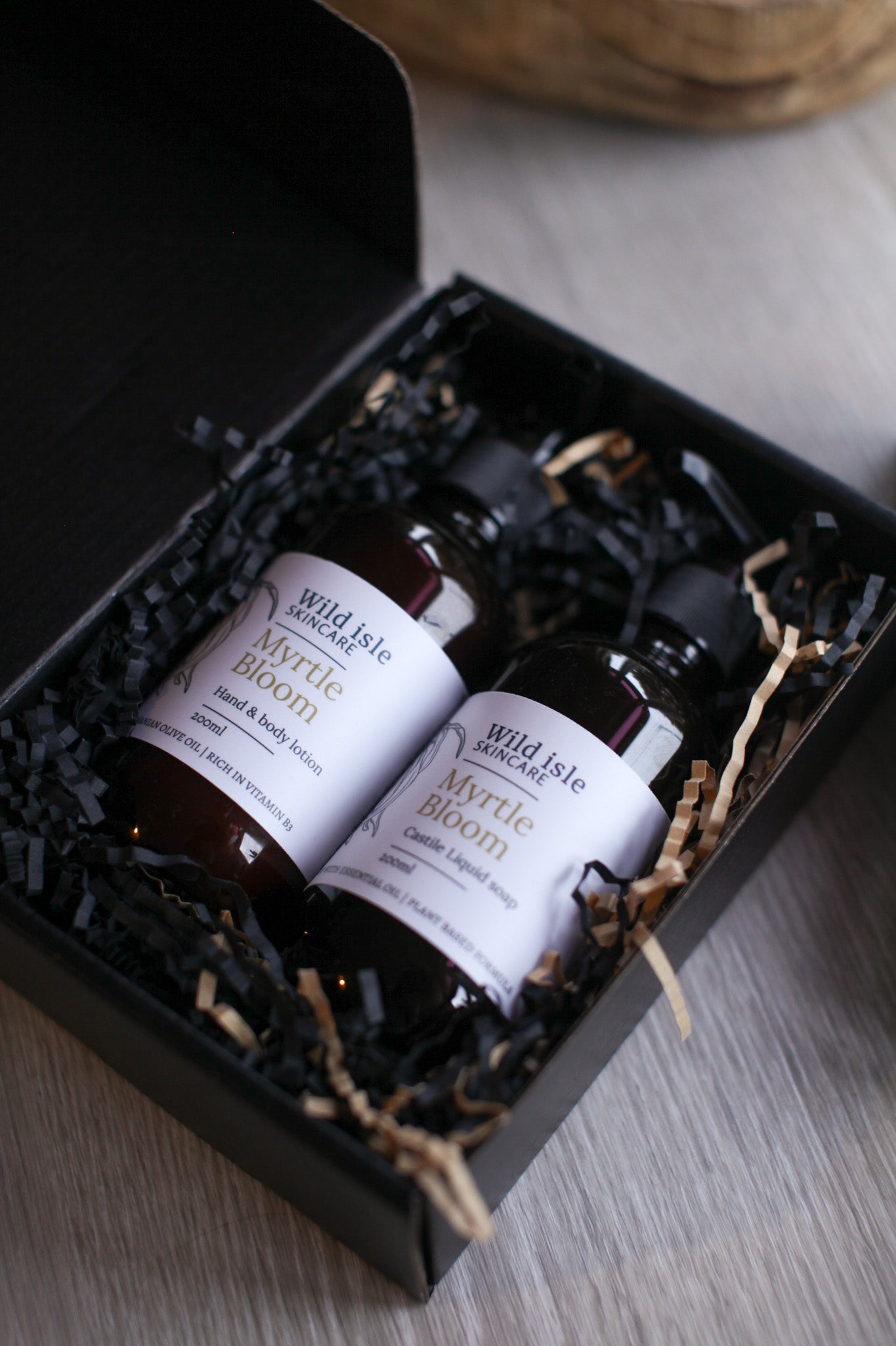 Three bottles of 'Myrtle Bloom' hand and body lotion in a black gift box on a wooden surface.